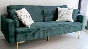 A green velvet sofa with wooden legs, two patterned pillows, and two framed pictures on a plain white wall. - Urbano Ñuñoa (Santiago)