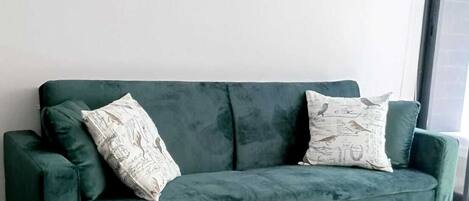 A green velvet sofa with wooden legs, two patterned pillows, and two framed pictures on a plain white wall.