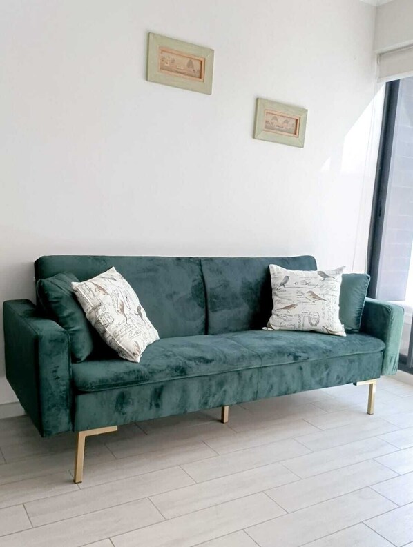 A green velvet sofa with wooden legs, two patterned pillows, and two framed pictures on a plain white wall.