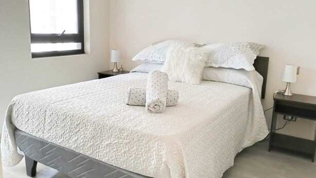 A neatly made bed with a textured white bedspread and pillows, two bedside lamps, and a window with blinds. - Urbano Ñuñoa (Santiago)