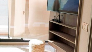 A modern living room with a large flat-screen TV mounted on a wooden wall, a fluffy white ottoman, and a sliding glass door leading to a balcony. - Urbano Ñuñoa (Santiago)