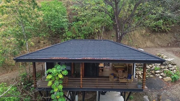 Une structure en bois dotée d’une terrasse et d’un toit sombre, entourée de verdure et bordée d’un chemin de terre.