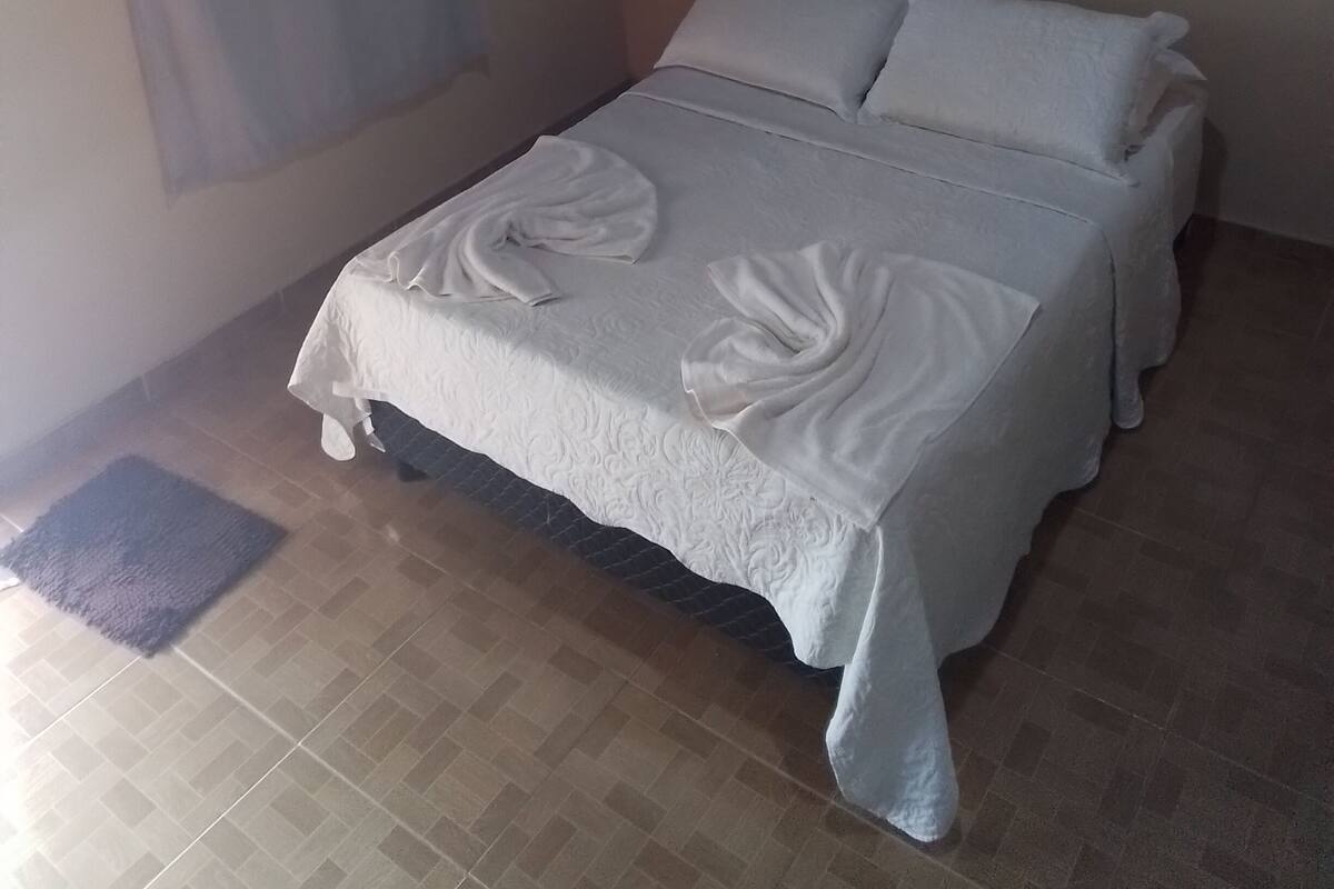 Un lit bien fait, avec du linge de lit et des serviettes blanches, une petite étagère au-dessus et une fenêtre avec un rideau.