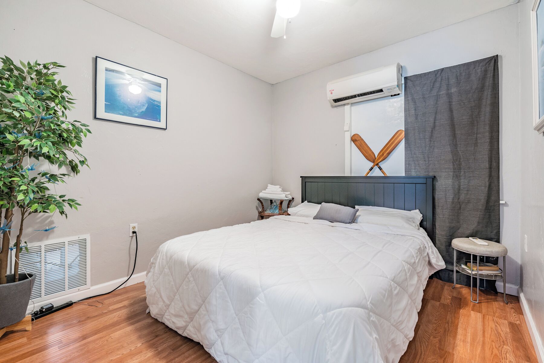 A neatly arranged bedroom with a bed, a potted plant, a wall-mounted air conditioner, and a framed picture.