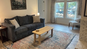 Una sala de estar con un sofá gris oscuro, una mesa de centro de madera, un tapete estampado y un sillón con cojín estampado.