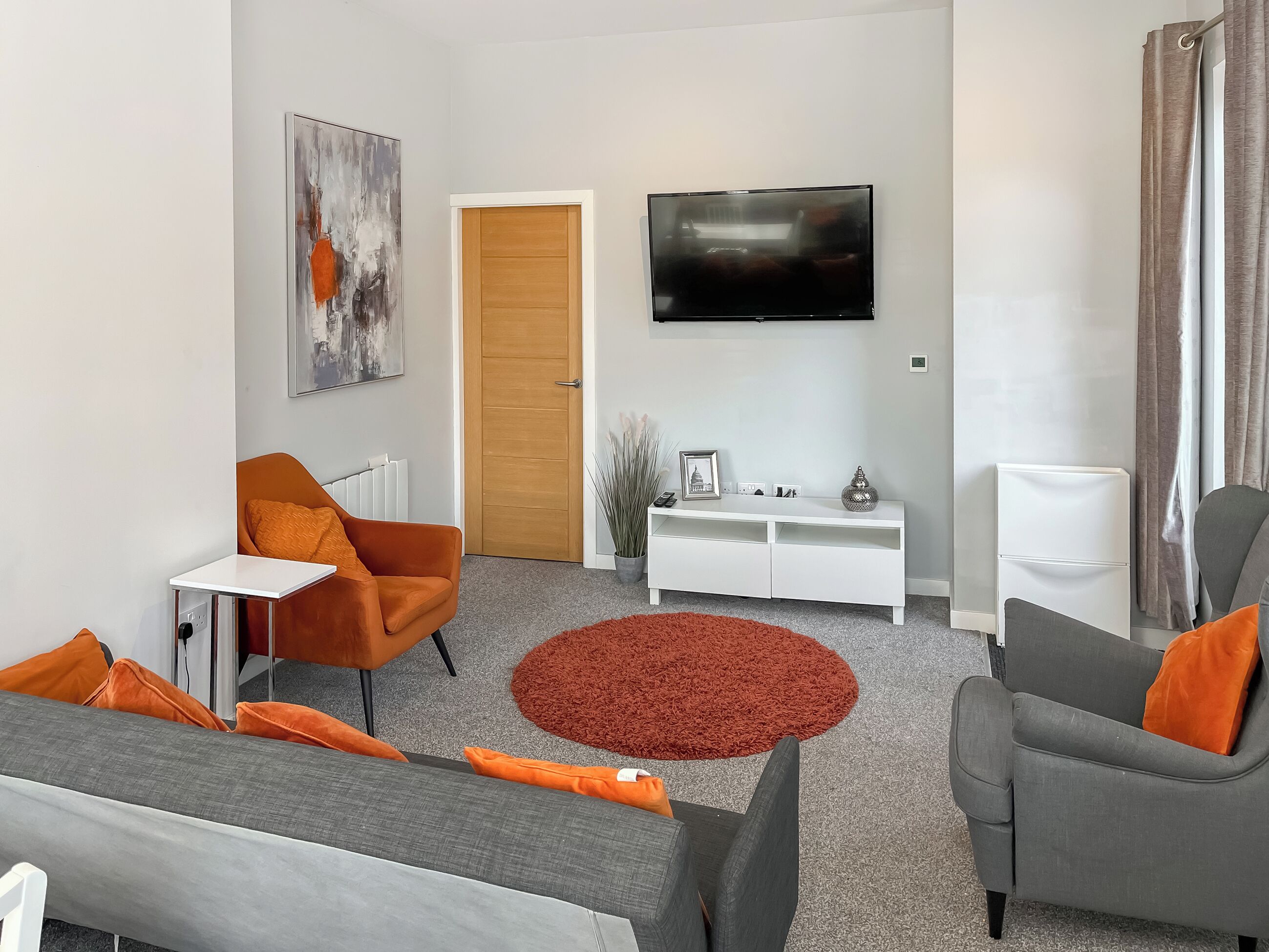 Ein modernes Wohnzimmer mit einem grauen Sofa, orangefarbenen Sesseln, einer weißen Unterhaltungsecke und einem flachbildfernseher an der Wand.