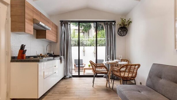 Eine kompakte Küche mit weißen Schränken, ein Essbereich mit Korbstühlen und ein graues Sofa.