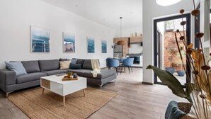 Ein modernes Wohnzimmer mit einem grauen Ecksofa, einem weißen Couchtisch und einem Essbereich mit blauen Stühlen. An der Wand befinden sich gerahmte Kunstwerke und ein großes Fenster sorgt für natürliches Licht.