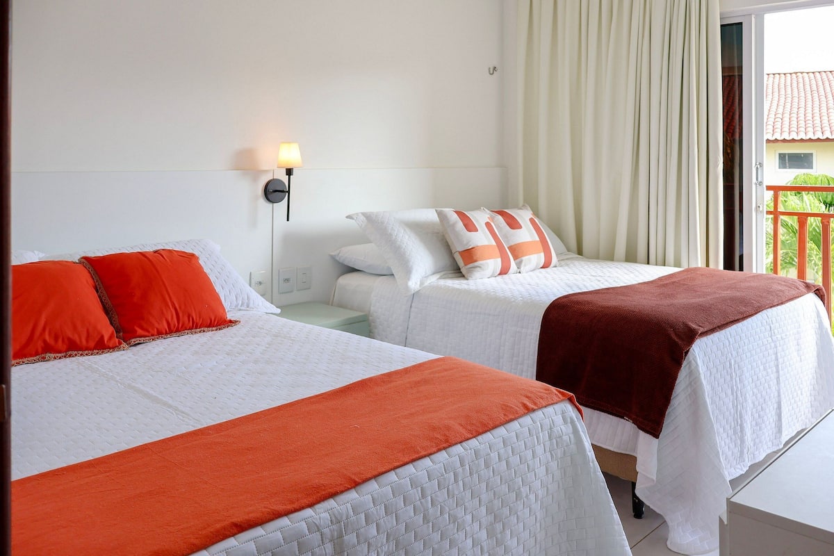 Two beds with white and orange bedding, a nightstand, a wall-mounted lamp, and a view of a balcony with a railing.