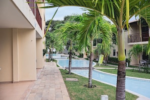 A pool area with palm trees, a tiled walkway, and a building with balconies. - Solar Água Pipa (Tibau do Sul)