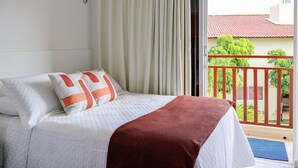 Habitación de hotel con cama, cortinas, balcón con barandilla y vistas a zonas verdes y edificios.