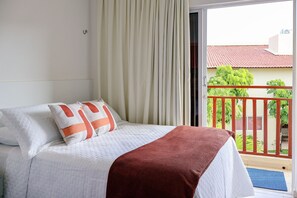 A hotel room with a bed, curtains, a balcony with a railing, and a view of greenery and buildings. - Solar Água Pipa (Tibau do Sul)