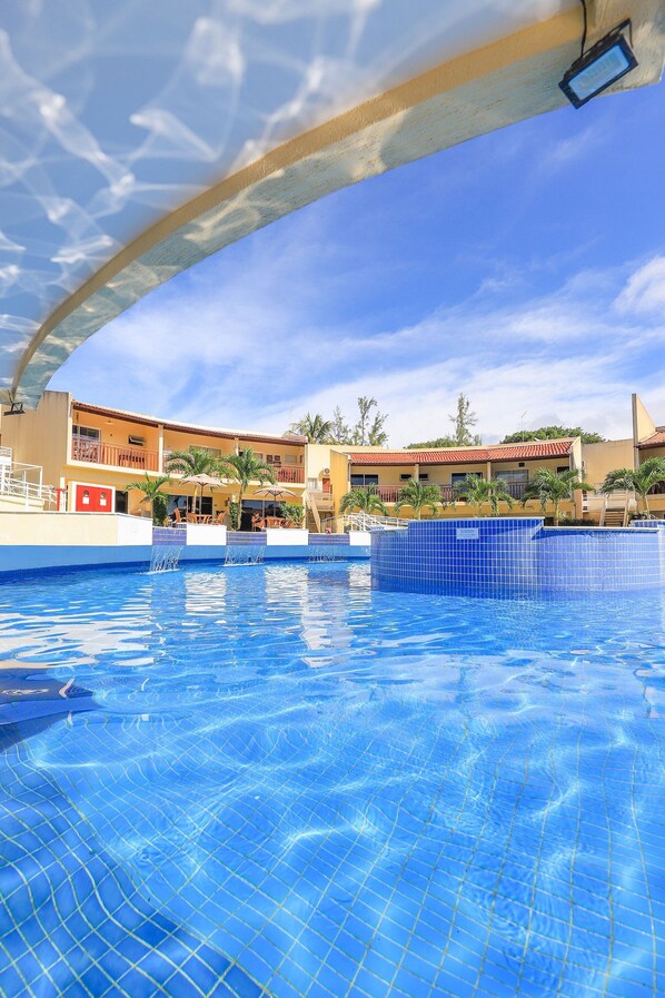 A swimming pool with clear blue water, surrounded by a building with balconies and palm trees. - Solar Água Pipa (Tibau do Sul)