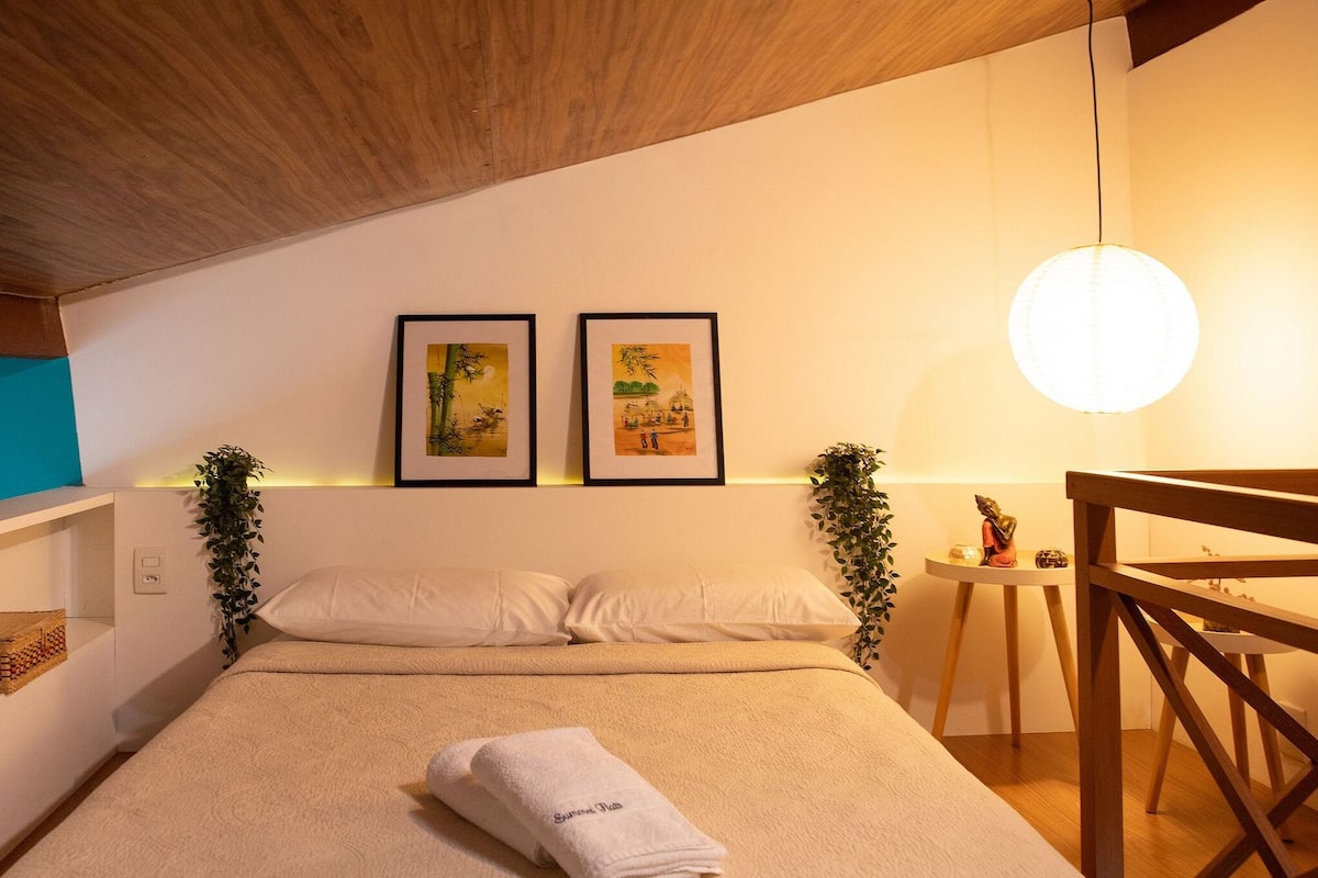 A bedroom with a bed, pillows, a bedside table, a lamp, and two framed artworks on the wall.