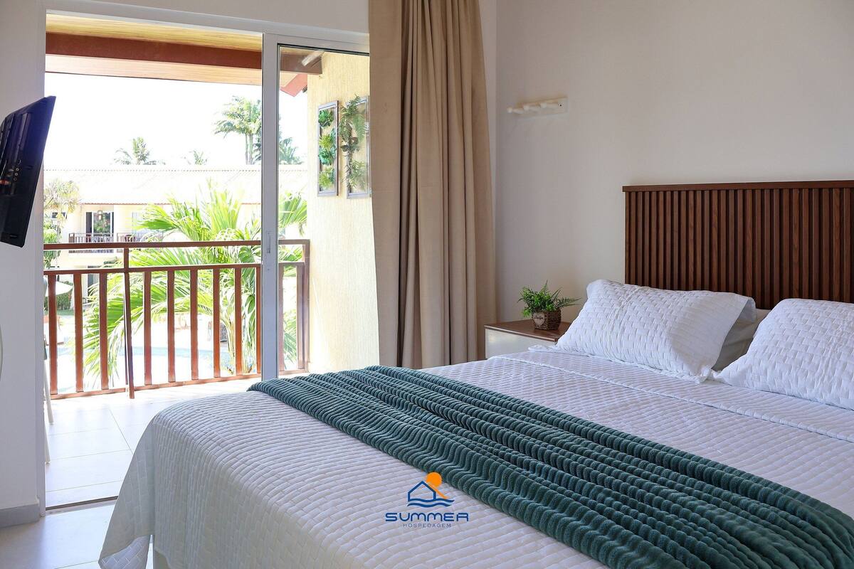 A hotel room with a bed, a nightstand, a TV, a balcony with plants, and a view of palm trees.