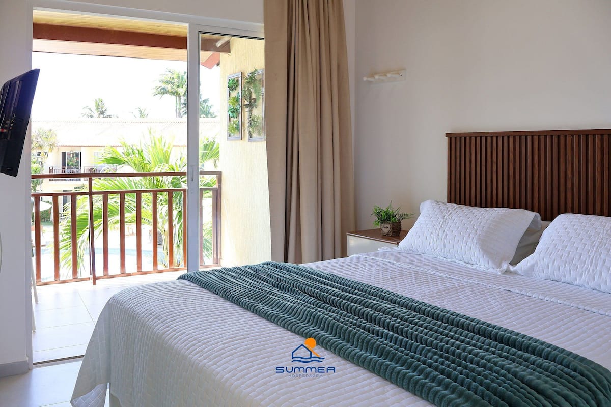 A hotel room with a bed, a nightstand, a TV, a balcony with plants, and a view of palm trees.