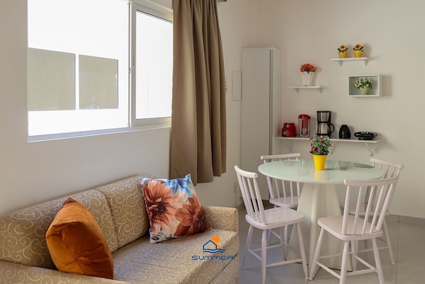 Un salon comprenant un canapé, une table en verre, des chaises blanches, une fenêtre avec des rideaux et un coin cuisine avec des étagères et des appareils électroménagers.