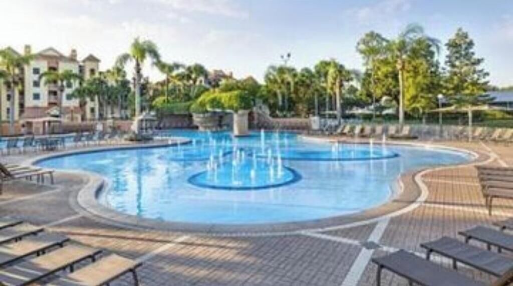 A large outdoor swimming pool with multiple fountains, surrounded by lounge chairs and a wooden deck.