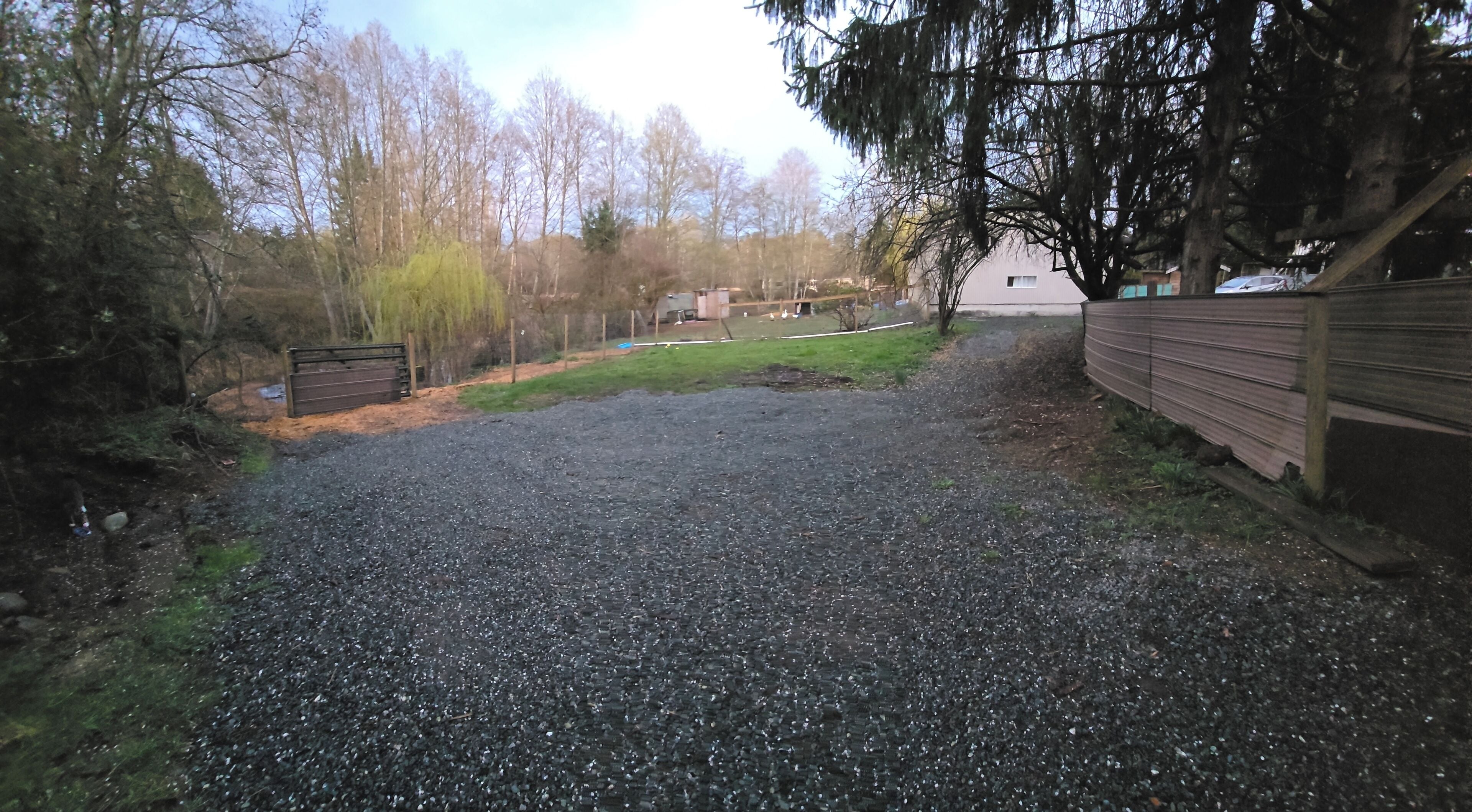 Een grindpad leidt naar een houten poort, omringd door bomen en een gazon.