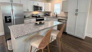A modern kitchen with white cabinets, a stainless steel refrigerator, a microwave, and a granite countertop. There are two wicker bar stools and a small island with a sink.
