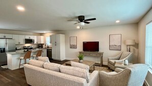 A modern living room with a sofa, chairs, a coffee table, and a TV. There is a kitchen area with cabinets and appliances in the background.