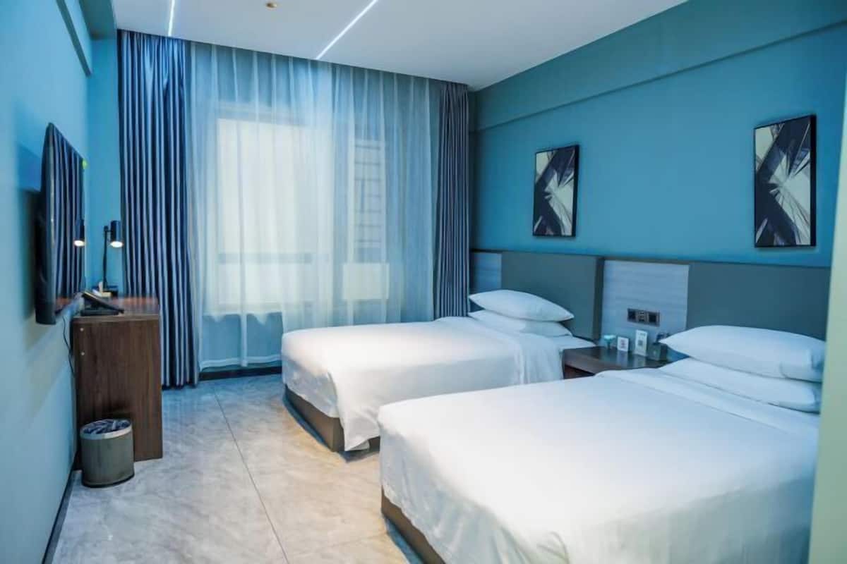 A hotel room with two beds, a desk, a trash bin, and artwork on the walls.