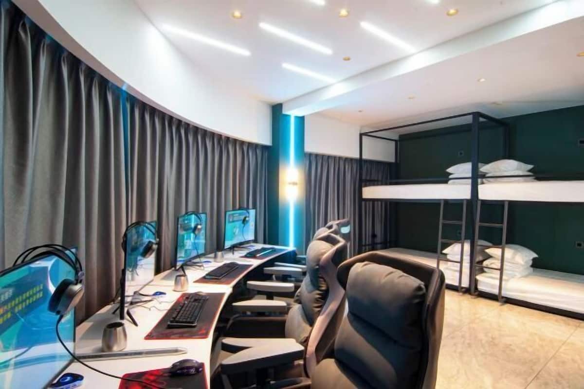A modern hotel room with a long desk, multiple monitors, a comfortable chair, a bunk bed, and a curved wall with curtains.