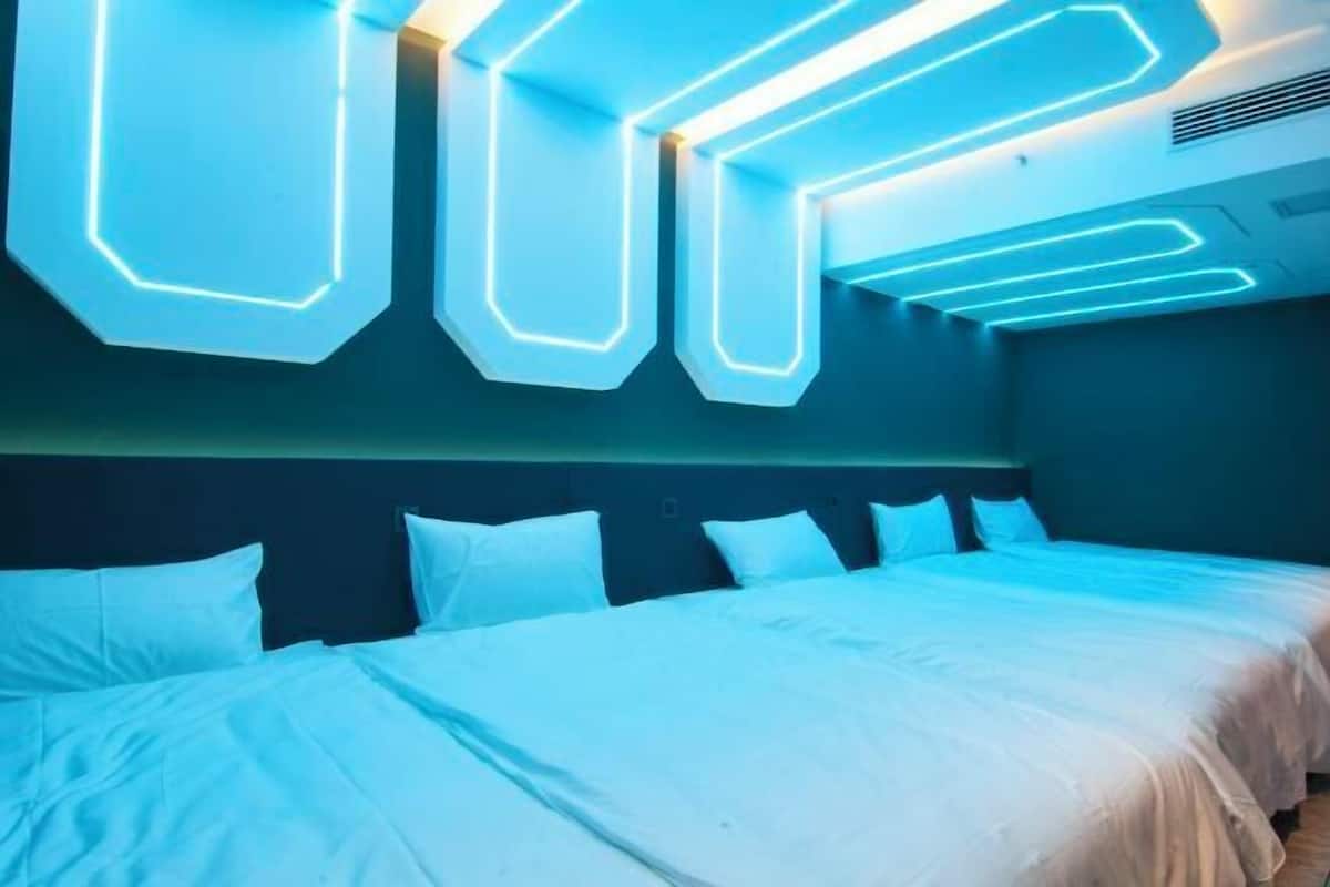 A modern hotel room with a large bed, a headboard with geometric light patterns, and a dark-colored wall.