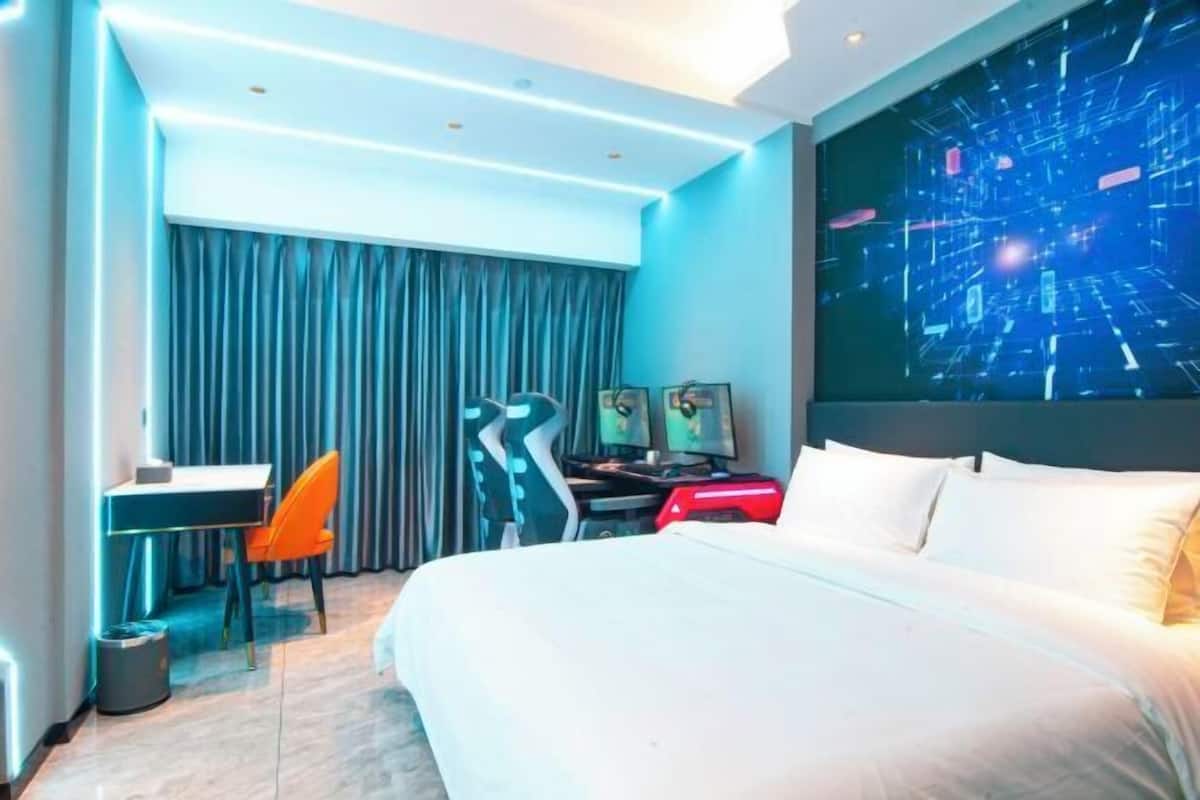A modern hotel room with a large bed, a desk with a chair, a computer, and a large digital art display on the wall.