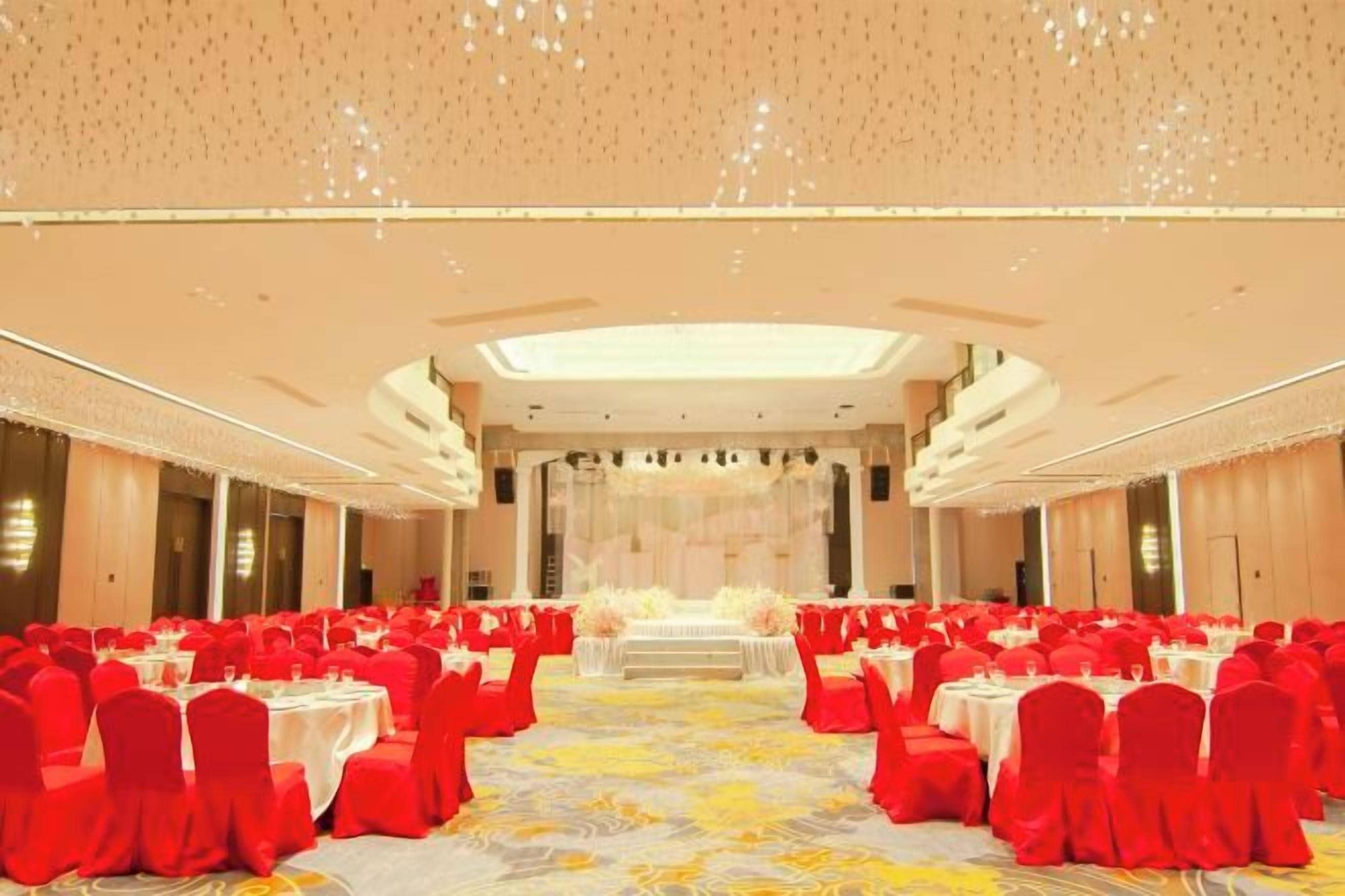 Un salón de banquetes con filas de mesas y sillas, un escenario central y una alfombra estampada.