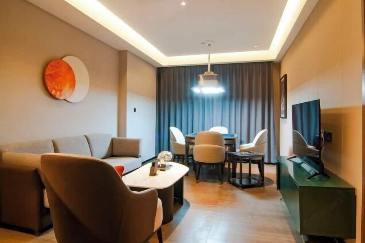 A modern hotel room with a sofa, a dining table with chairs, a TV, and a large window with curtains.