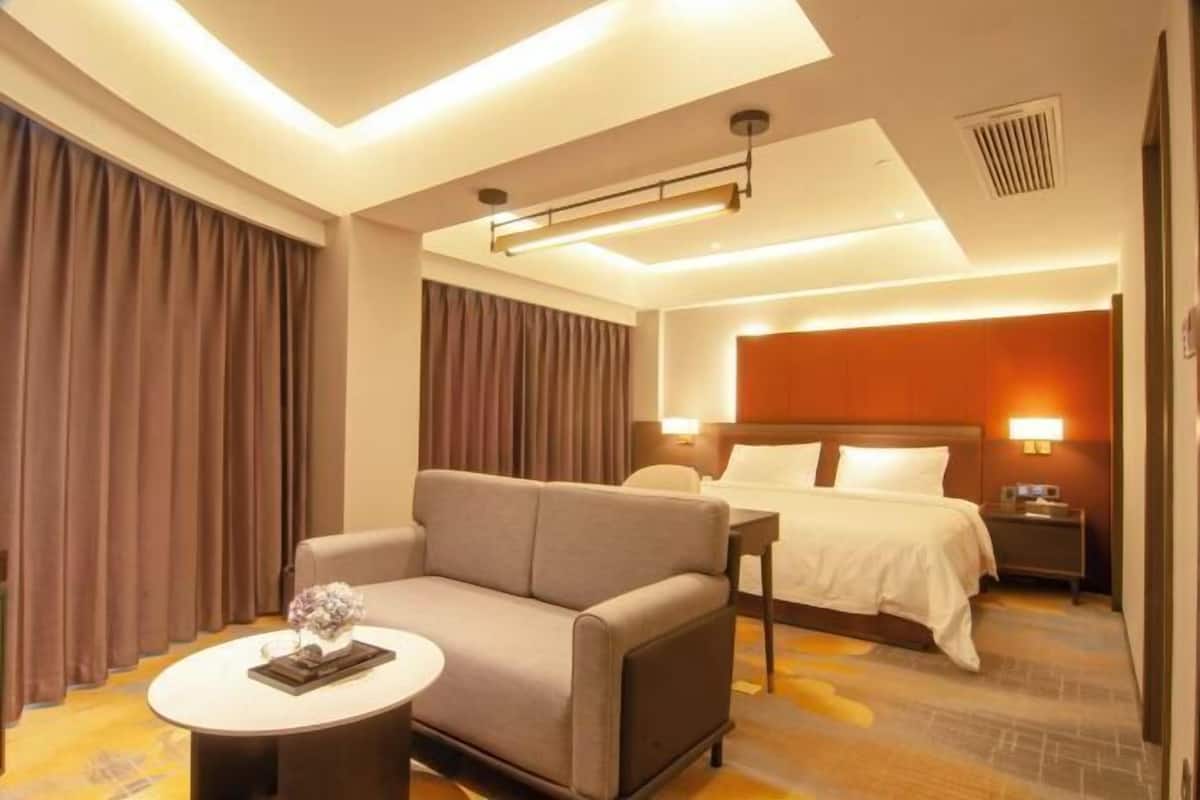 A hotel room with a sofa, a small round table with a vase, a bed with a headboard, and a nightstand with a lamp.
