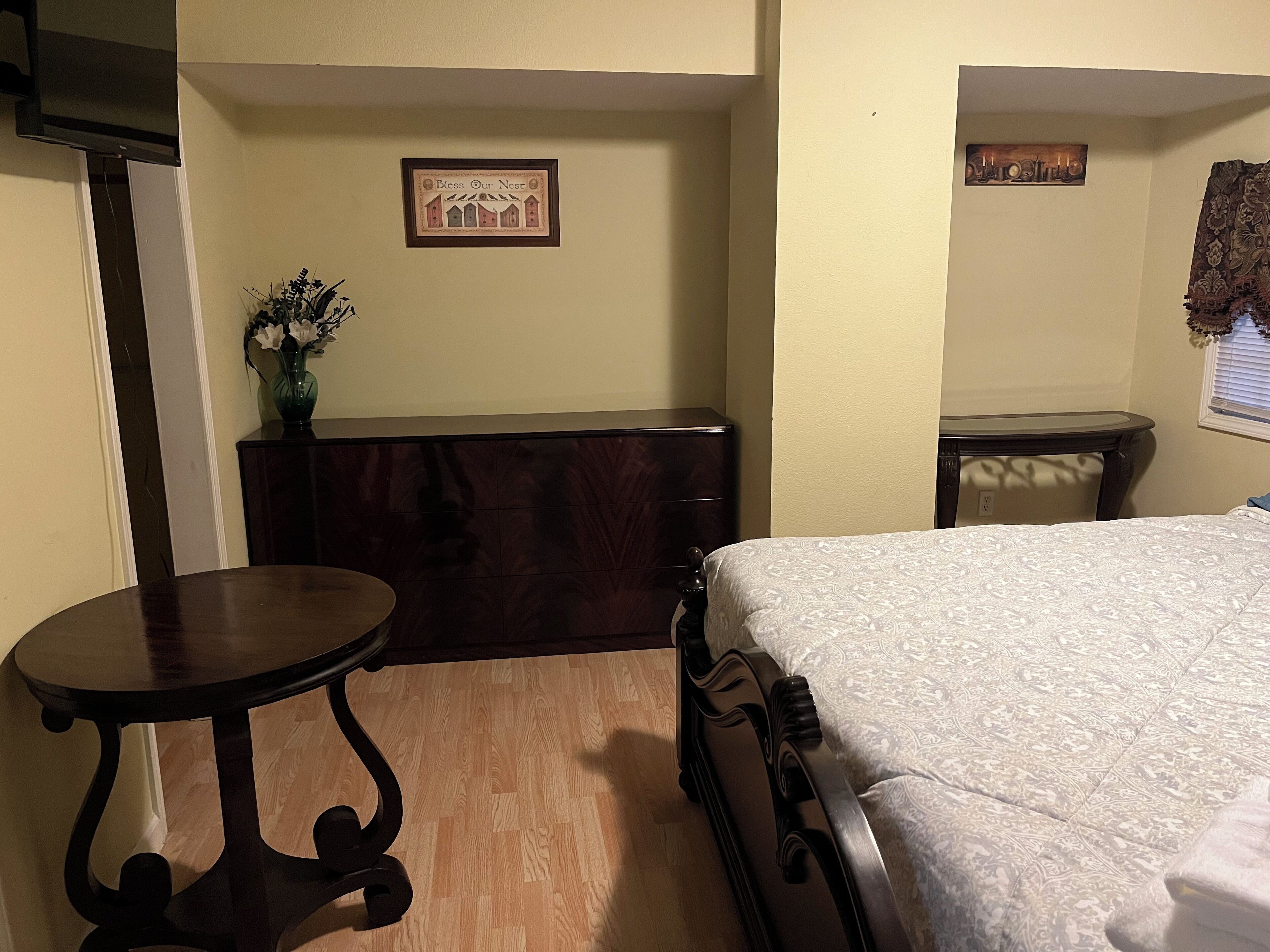 A bedroom with a bed, a dresser, a small table, and a wall-mounted shelf with a framed picture.