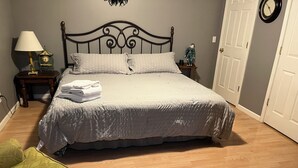 Una cama bien tendida con edredón blanco y almohadas, suelo de madera, un sillón verde, una mesita de noche con una lámpara y un reloj de pared.