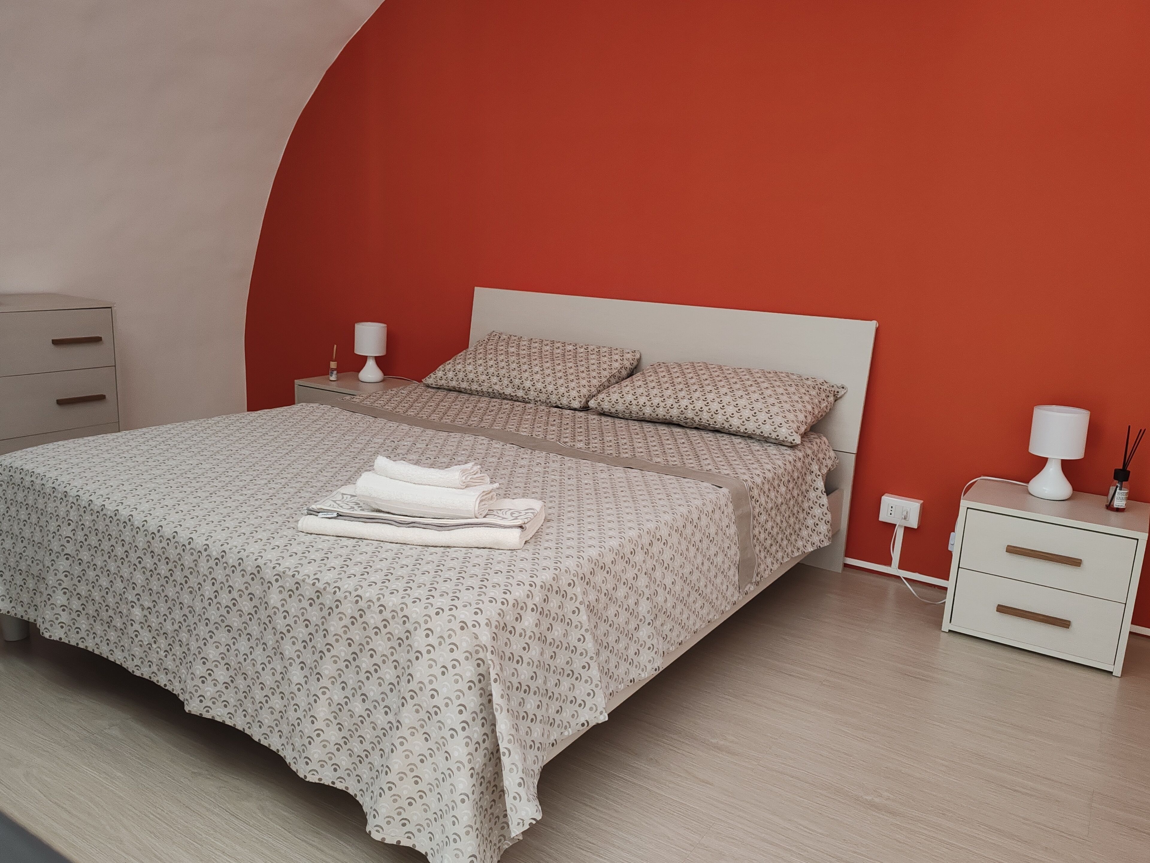 A neatly made bed with a patterned bedspread and pillows, a bedside table with a lamp, and a drawer unit.