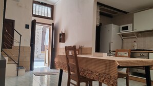Een eetgedeelte met een geruit tafelkleed, houten stoelen, een keuken met een magnetron en fornuis, en een trap naar een buitenruimte.
