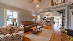 A spacious living room with a sofa, coffee table, and patterned chairs. A kitchen is visible in the background with a dining area. - Outer Banks Golf Cottage (Powells Point)