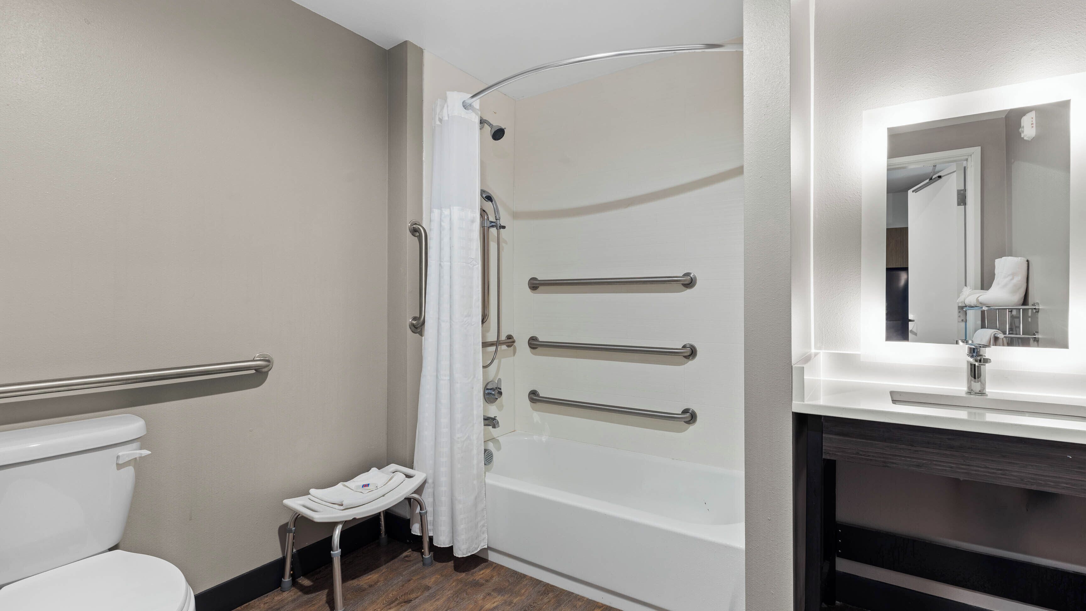 A bathroom with a white toilet, a shower with grab bars, a small step stool, and a mirror above the sink.
