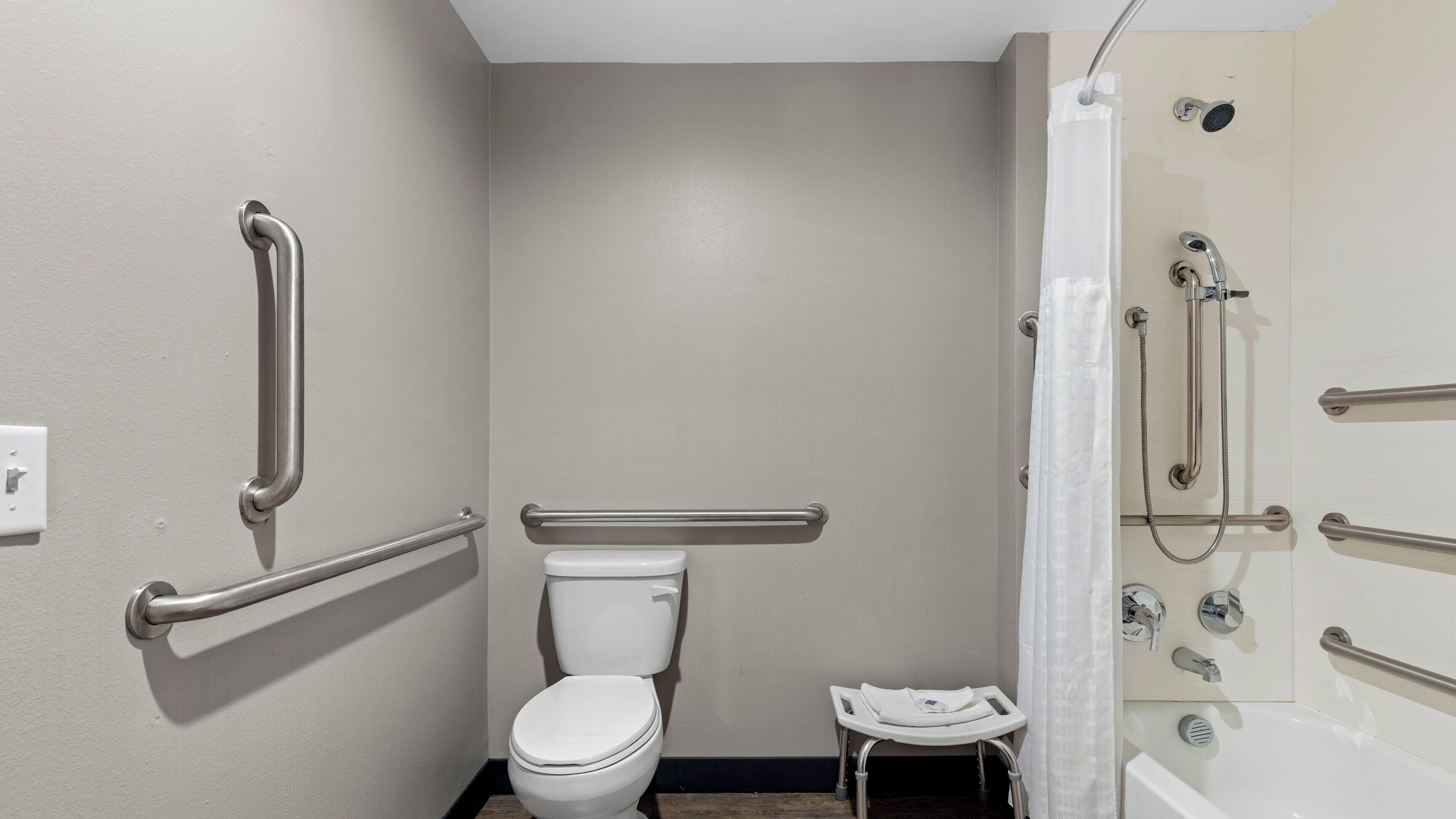 Un baño con inodoro, ducha con barras de apoyo y un pequeño taburete.
