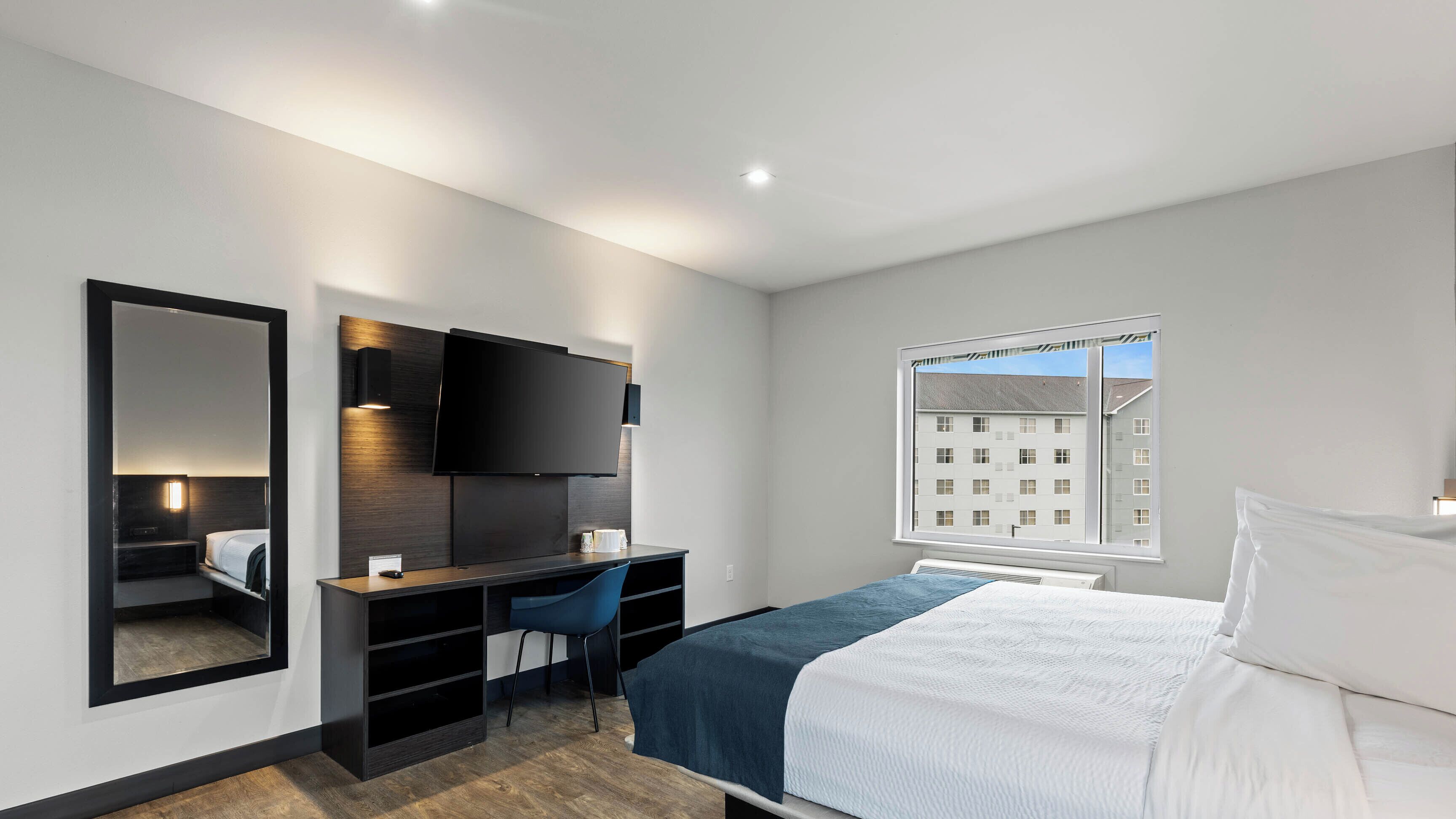 A modern hotel room with a bed, a desk with a chair, a flat-screen TV mounted on the wall, and a window with a view of buildings.