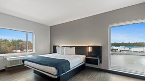 Habitación de hotel moderna con una cama grande, una mesita de noche, una lámpara de pared y vistas a una masa de agua y edificios industriales.