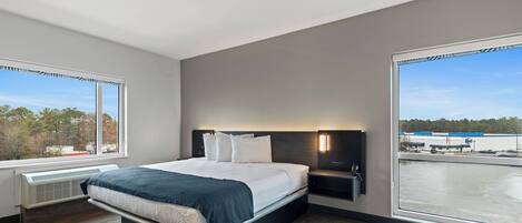 A modern hotel room with a large bed, a nightstand, a wall-mounted lamp, and a view of a body of water and industrial buildings.