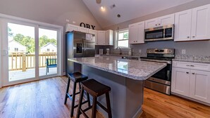 A modern kitchen with white cabinets, a large island with stools, stainless steel appliances, and a sliding glass door leading to an outdoor deck.