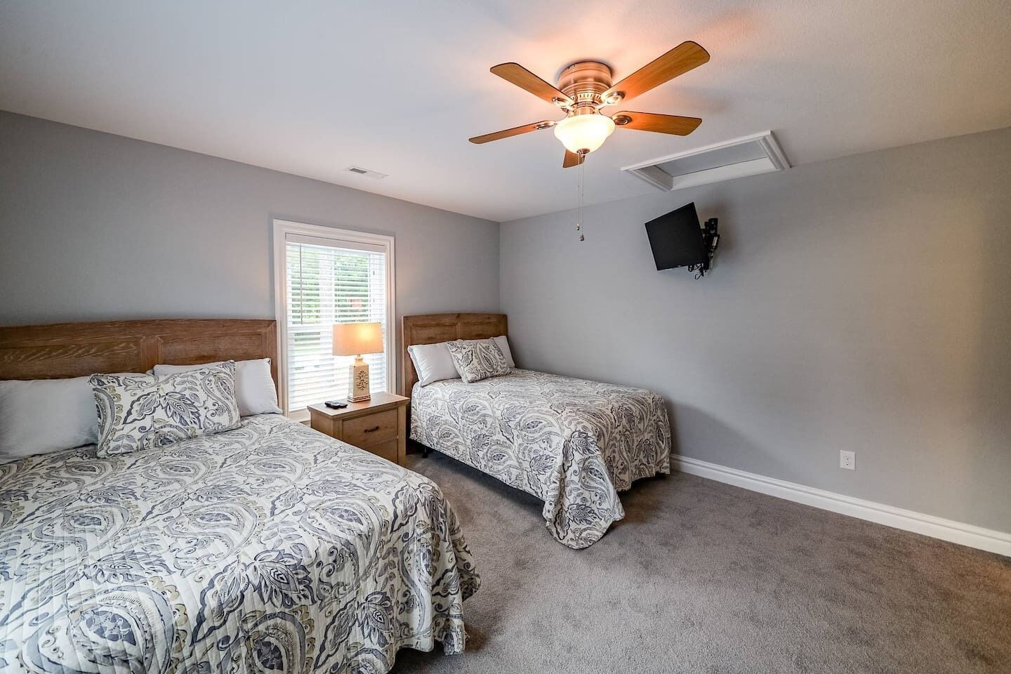 A bedroom with two beds, a ceiling fan, a window with blinds, and a mounted TV.