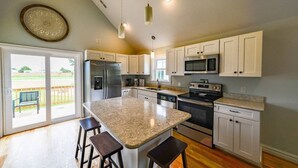 A spacious kitchen with a large island, stainless steel appliances, and a sliding glass door leading to a patio. - OBX Cottages The Snead Cottage (Powells Point)
