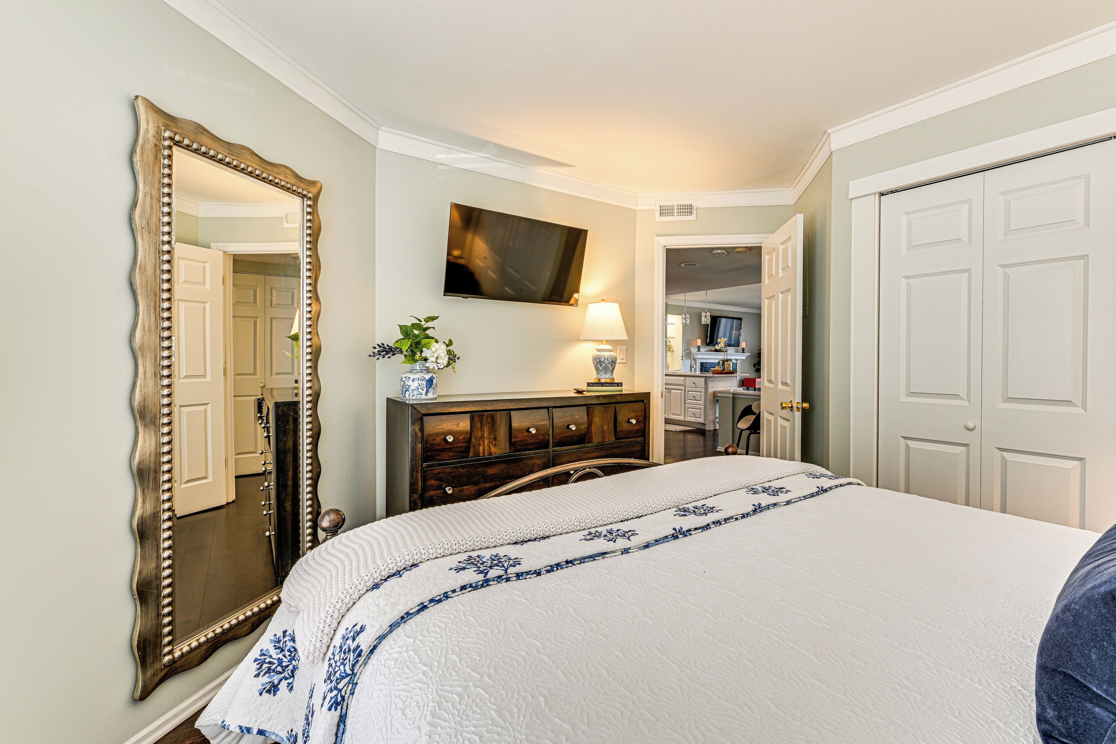 A bedroom with a large bed, a dresser, a mirror, a television, and a door leading to another room.