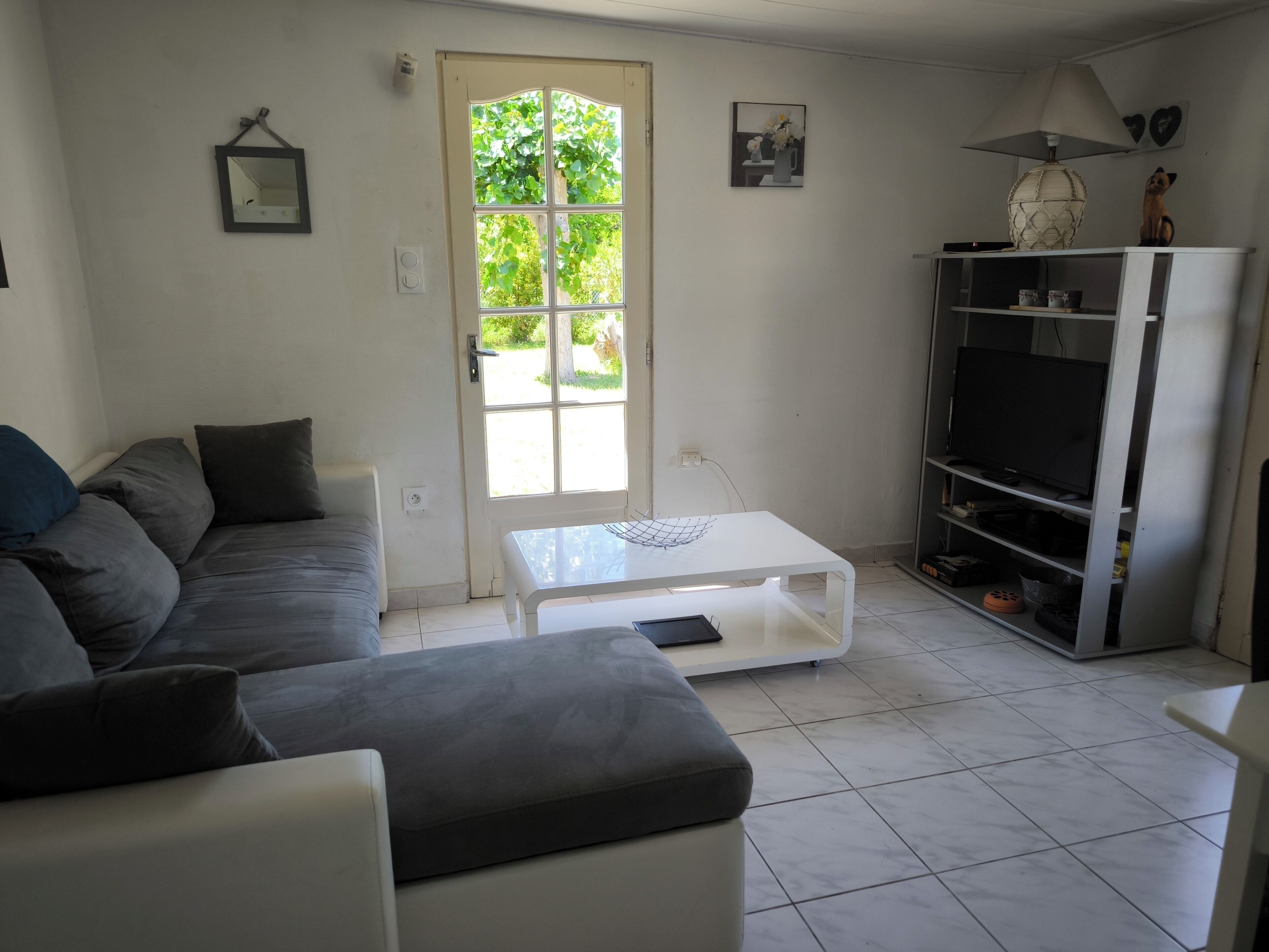 A living room with a grey sofa, a white coffee table, a TV stand with a TV, and a door leading outside.