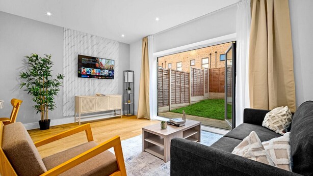 Een moderne woonkamer met een televisie, een bank, een fauteuil, een salontafel en uitzicht op een bakstenen muur en tuin.