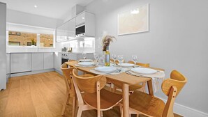 Een moderne keuken met witte kasten, een houten eettafel voor vier personen en een vaas met bloemen aan de muur.