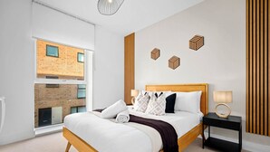 Een moderne slaapkamer met een groot bed, een nachtkastje, een lamp en uitzicht op een bakstenen gebouw door een groot raam.